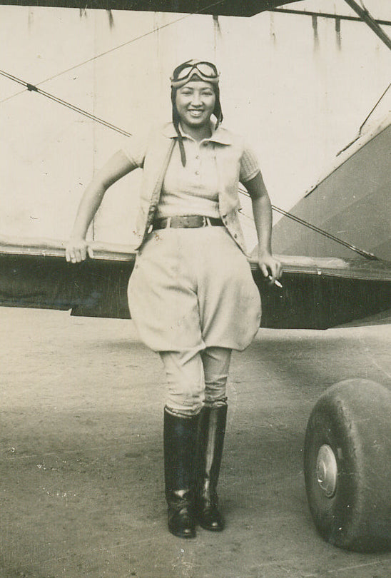 A vintage black and white photograph of a smiling woman in an early 20th-century flying outfit, including aviator goggles, a short-sleeve collared shirt, a skirt, and tall boots, standing in front of an airplane on an airfield.