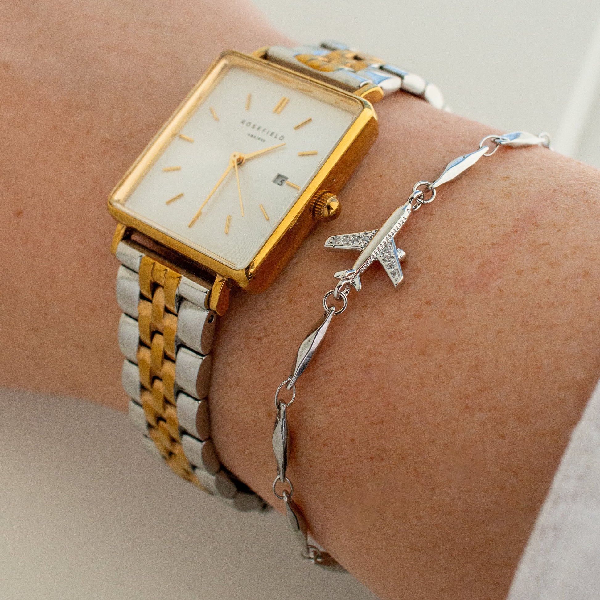A close-up of a person's wrist wearing a gold and silver two-tone rectangular watch with a white dial, layered with a silver chain bracelet featuring a small airplane charm.
