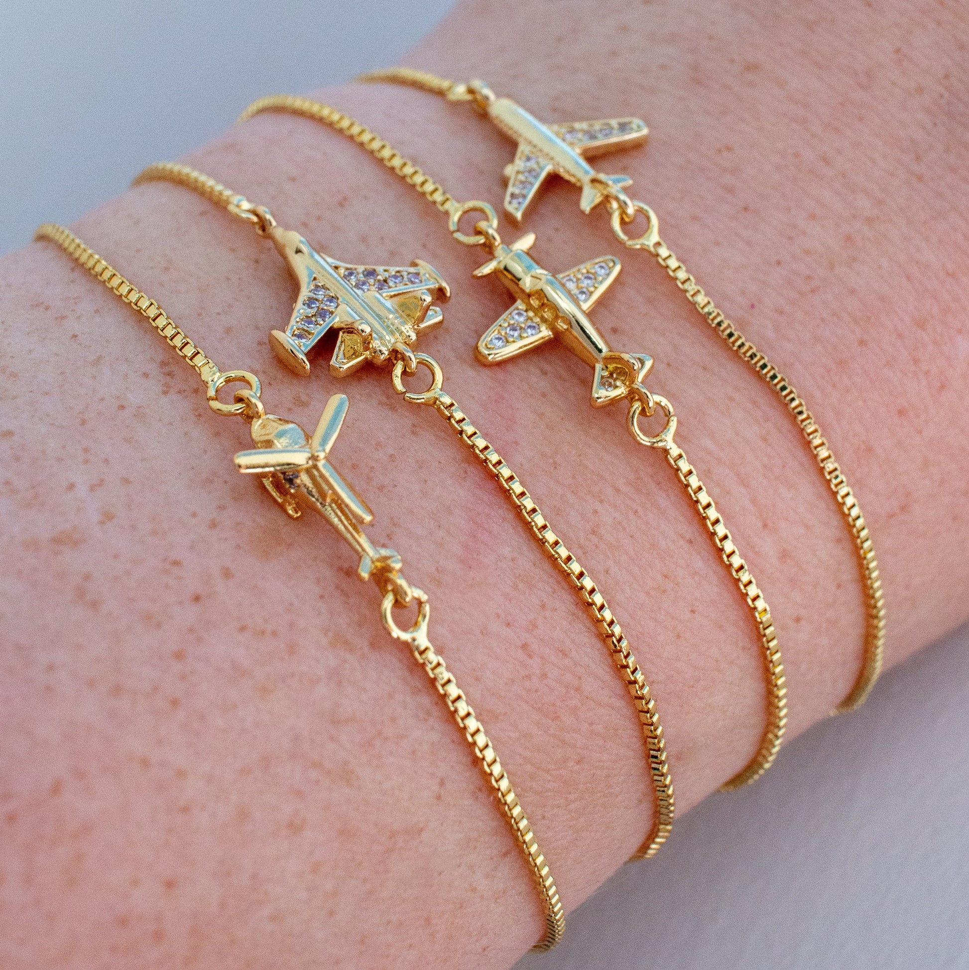 A close-up of a person's wrist wearing four delicate gold chain bracelets, each adorned with a small airplane charm.