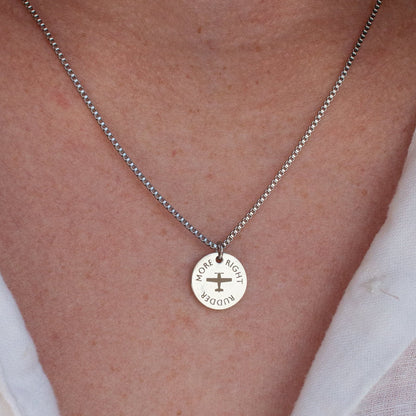 A close-up of a person wearing a delicate silver chain necklace with a round silver pendant featuring a cross and the words "more right rudder" engraved, styled with a white collared shirt.