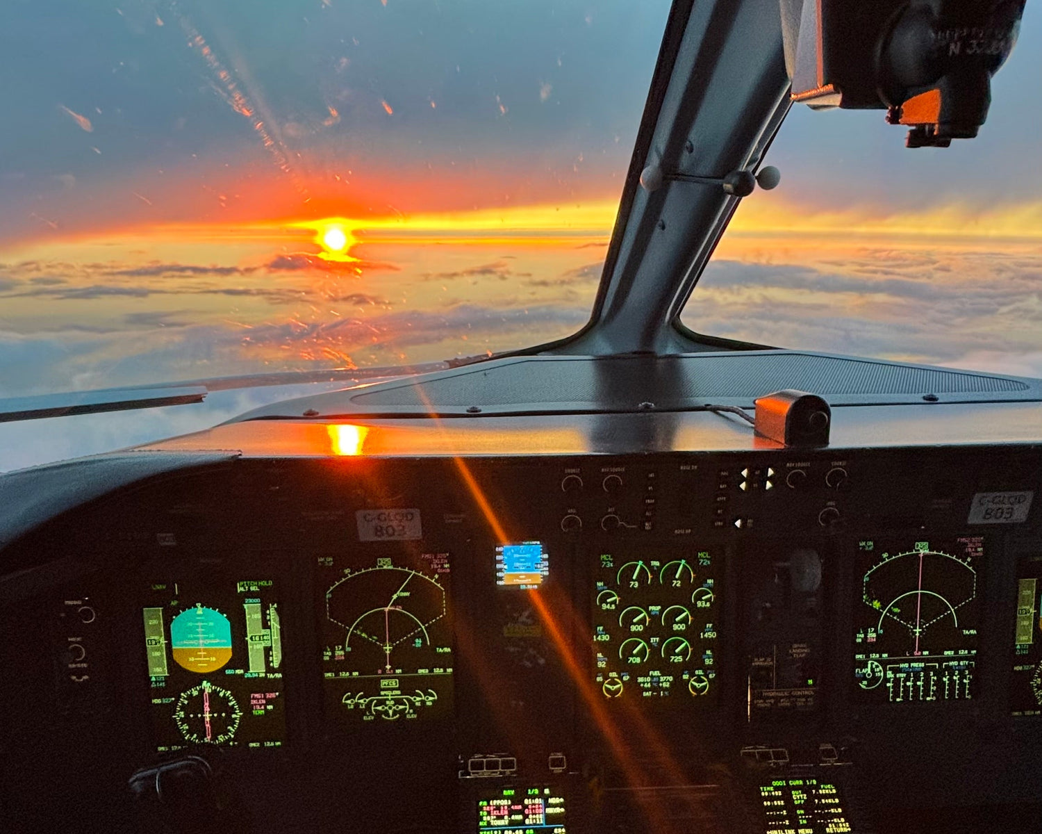 View from inside an airplane cockpit looking out over a sunset over clouds, with illuminated instrument panels below.