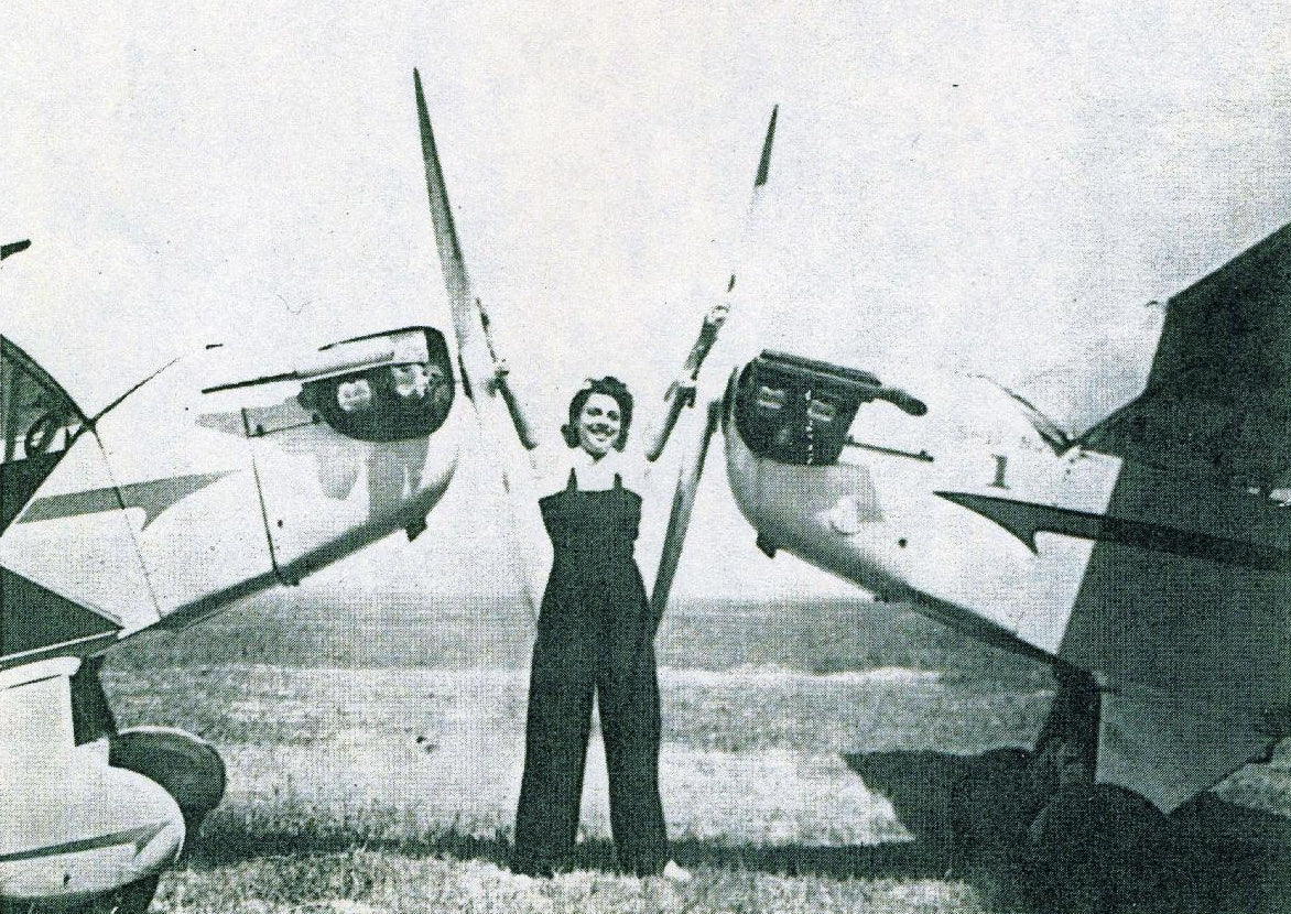 Black and white vintage photograph of a woman standing between two small aircraft on an airfield, wearing dark overalls.